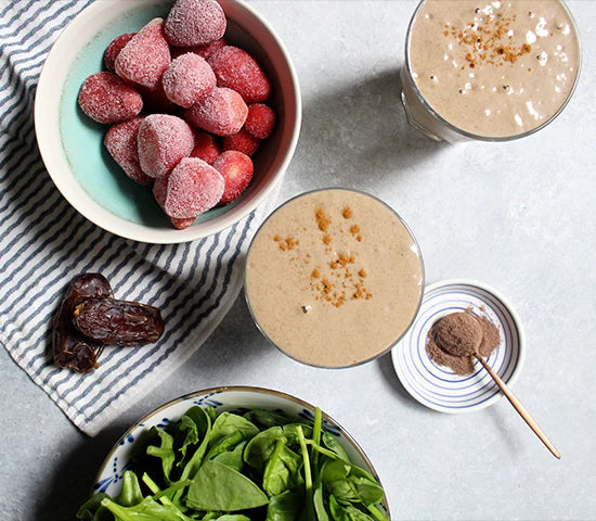 Flat lay with bowl of strawberries, spinach, and loose cacao powder around two glasses of strawberry cacao shake