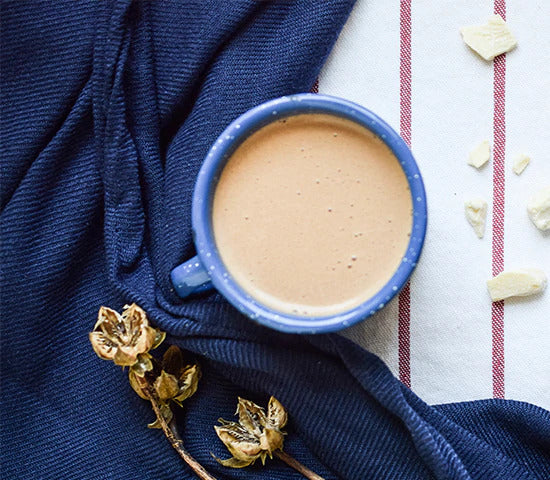 A blue ceramic mug filled with a cacao butter mocha made with Navitas Organics Cacao Butter Wafers and Cacao Powder, surrounded by a blue tablecloth and dried flowers