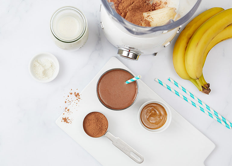 A chocolate smoothie made with Navitas Organics Cacao Powder and Maca Powder in a glass on a cutting board with a scoop of cacao powder, nut butter and a glass of plant-based milk