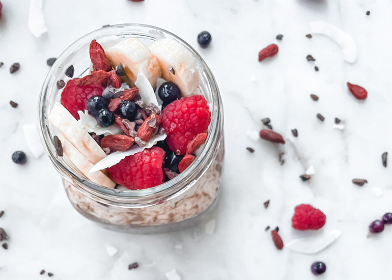 A mason jar filled with overnight oats made with Navitas Organics Acai Powder, Cacao+ Antioxidant Blend and Chia Seeds, topped with berries, coconut flakes and banana slices