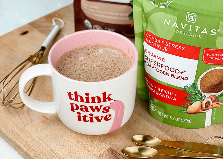 A mug filled with hot cocoa made with Navitas Organics Cacao Powder and Superfood+ Adaptogen Blend on a wooden cutting board with a whisk and measuring spoons