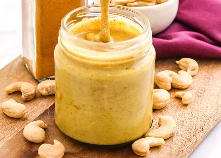 A jar of golden milk cashew butter made with Navitas Organics Cashew Nuts and Turmeric Powder on a cutting board surrounded by cashews