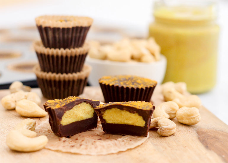 Stacks of chocolate golden milk cashew butter cups made with Navitas Organics Cacao Butter, Cacao Powder, Cashew Nuts and Turmeric Powder on a wooden cutting board surrounded by cashews