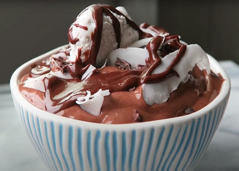 A bowl of chocolate nice cream made with Navitas Organics Cacao+ Longevity Blend, topped with coconut whipped cream, shredded coconut, nut butter and Navitas Organics Cacao Nibs