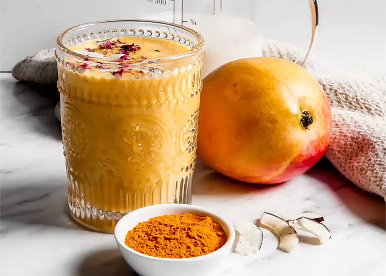 A glass filled with a mango turmeric smoothie made with Navitas Organics Turmeric Powder and Chia Seeds, alongside a mango fruit and a dish filled with turmeric powder