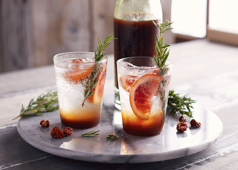Two glasses filled with a goldenberry shrub made with Navitas Organics Goldenberries, garnished with oranges and rosemary