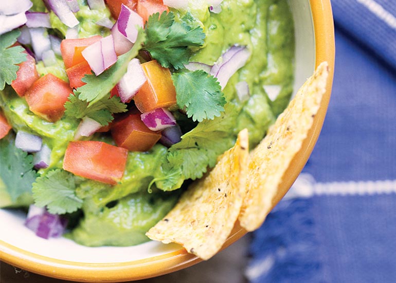 A bowl of superfood guacamole made with Navitas Organics Wheatgrass Powder, garnished with chopped tomatoes, red onions and cilantro, with a side of tortilla chips