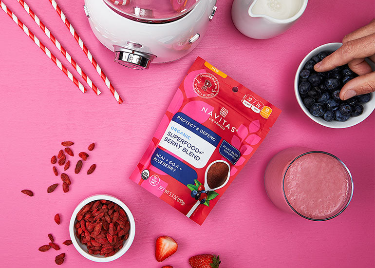 A pink smoothie made with Navitas Organics Superfood+ Berry Blend next to a dish filled with blueberries and a dish filled with Navitas Organics Goji Berries
