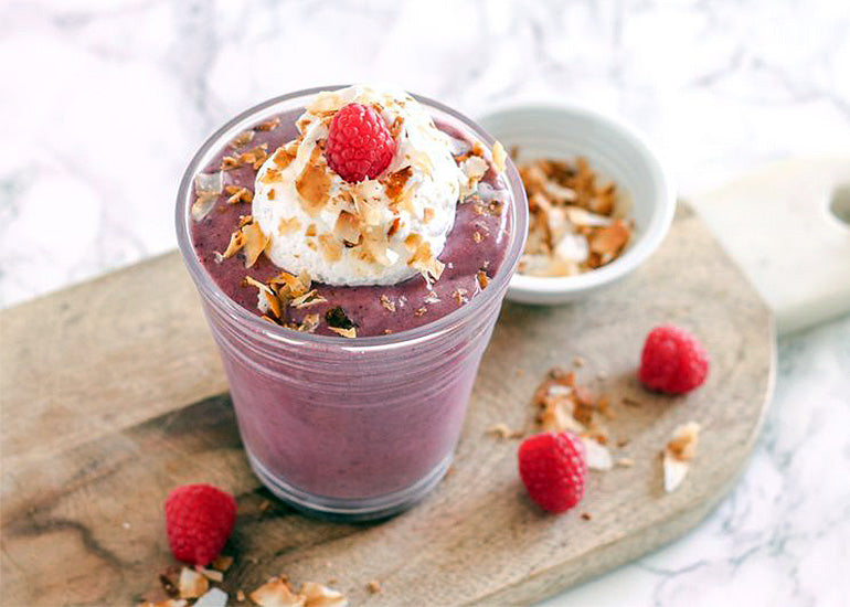 A glass filled with a pink smoothie made with Navitas Organics Hemp Seeds and Superfood+ Berry Blend, topped with whipped cream, toasted coconut and fresh raspberries