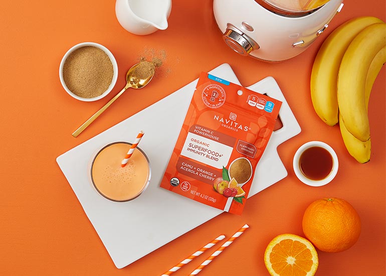 An orange smoothie made with Navitas Organics Superfood+ Immunity Blend on a cutting board alongside oranges and bananas
