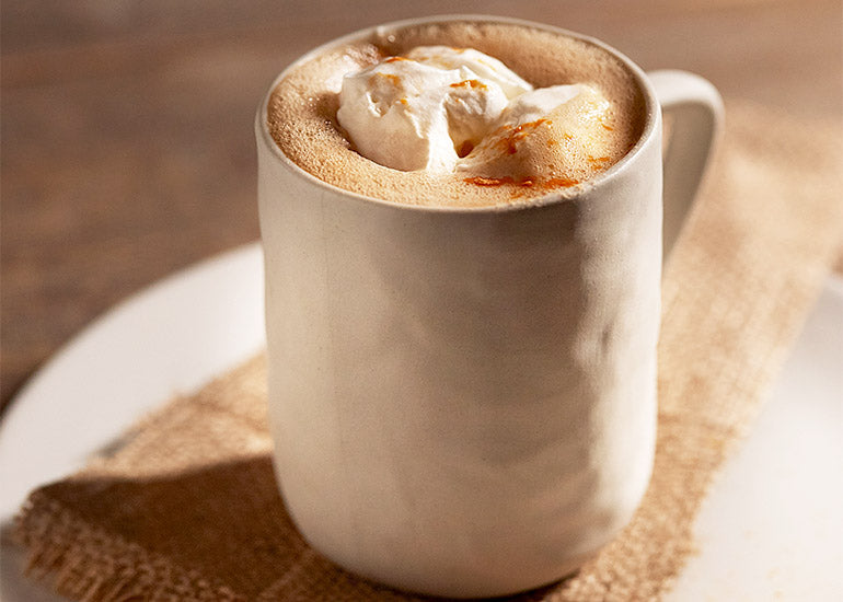 A mug filled with a foamy latte made with Navitas Organics Cacao Butter and Goji Berry Powder, topped with whipped cream and cinnamon
