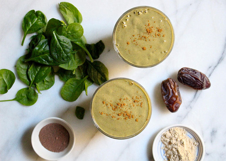 Two green smoothies made with Navitas Organics Cacao & Greens Essential Blend, Superfood+ Adaptogen Blend and Chia Seeds, topped with cinnamon, surrounded by fresh spinach and dates