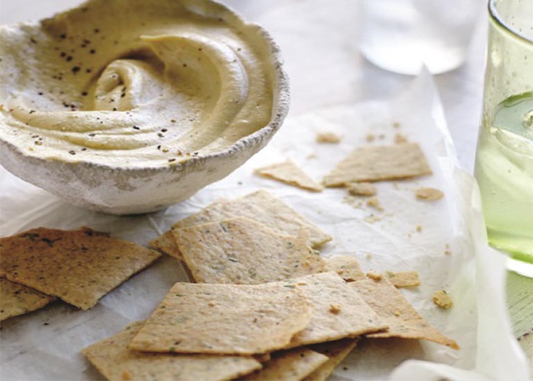 A pile of almond crackers made with Navitas Organics Hemp Seeds, next to a bowl of hummus for dipping.
