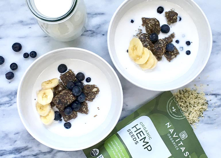 Two breakfast bowls made with Navitas Organics Blueberry Hemp Power Snacks and Hemp Seeds, topped with banana slices and blueberries.