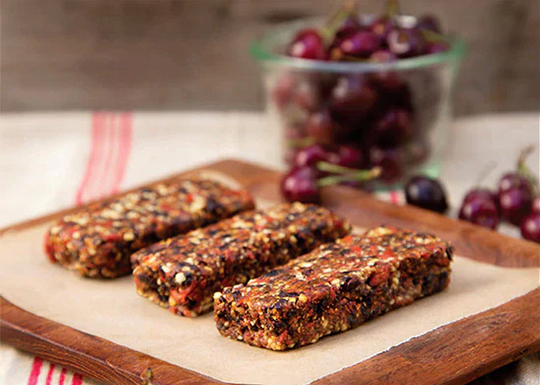 A row of chocolate cherry goji bars made with Navitas Organics Goji Berries and Cacao Nibs on a cutting board surrounded by fresh cherries.