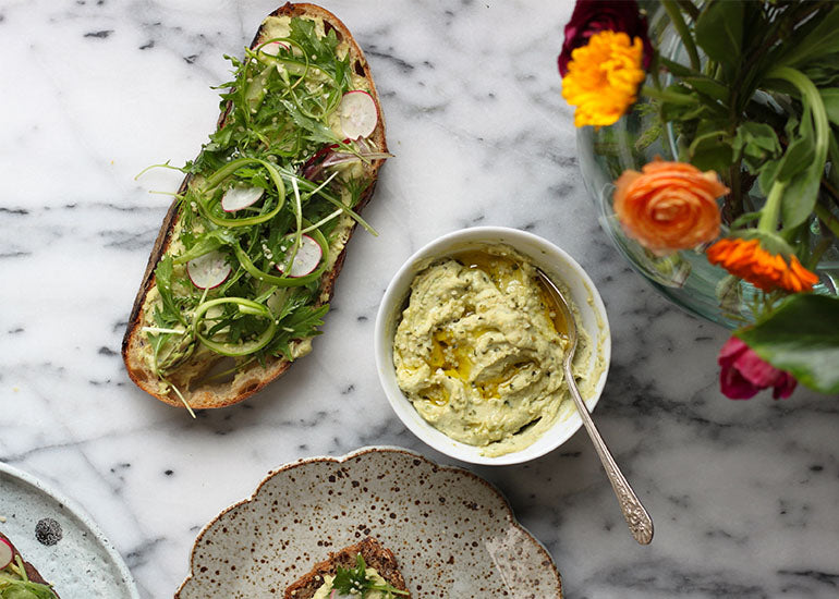 A slice of toast spread with hummus made with Navitas Organics Hemp Seeds, topped with fresh herbs, greens and sliced radishes.