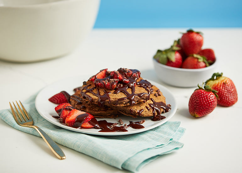 A plate of chocolate-oat protein pancakes made with Navitas Organics Cacao Powder, Chia Powder and Cacao Nibs, drizzled with chocolate sauce and topped with fresh strawberries.
