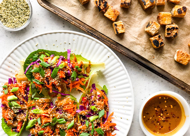 A plate of tofu lettuce wraps with maca peanut sauce made with Navitas Organics Maca Powder next to a pan of crispy tofu and a dish filled with Navitas Organics Hemp Seeds
