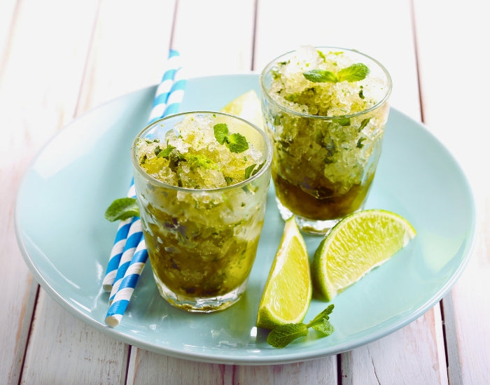 Two lime granitas made with Navitas Organics Superfood+ Greens Blend in glasses on a blue plate with fresh limes and straws.