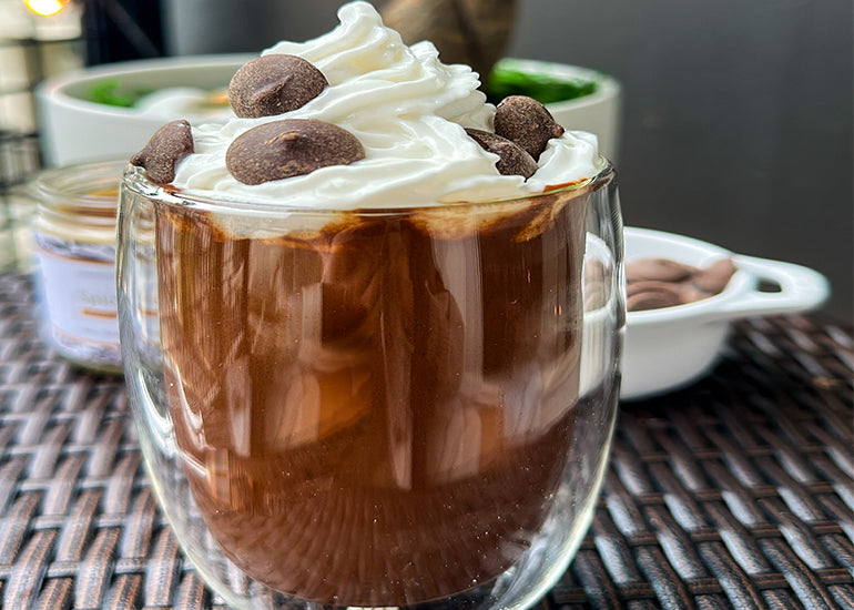 A glass filled with a French hot chocolate made with Navitas Organics Bittersweet Cacao Wafers, topped with dairy-free whipped cream and more cacao wafers.