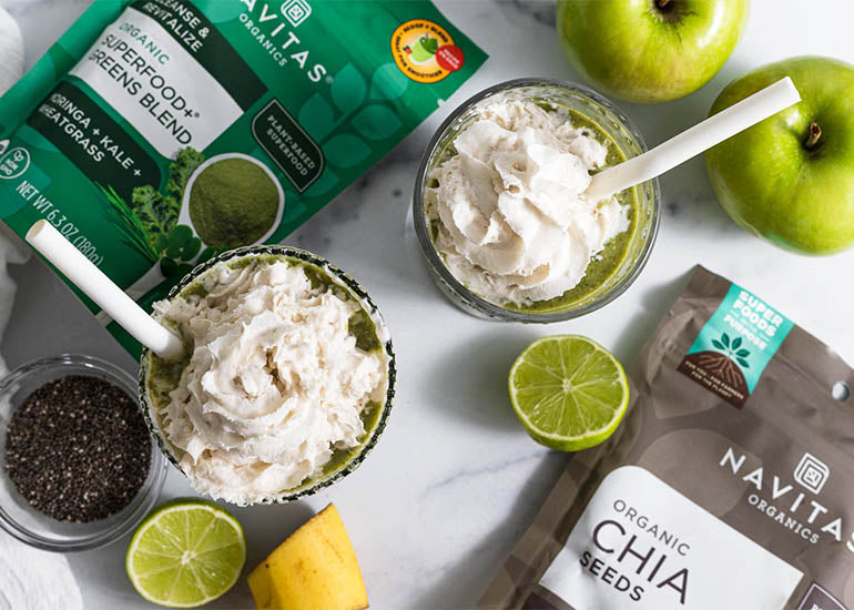 Two green smoothies made with Navitas Organics Superfood+ Greens Blend and Chia Seeds, topped with dairy-free whipped cream