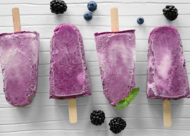 Four purple frozen popsicles sitting on a wooden table surrounded by fresh blueberries and blackberries