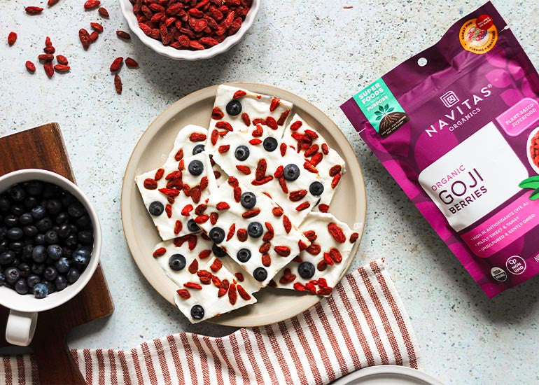 A plate of goji blueberry frozen yogurt bark pieces surrounded by dishes filled with fresh blueberries and Navitas Organics Goji Berries