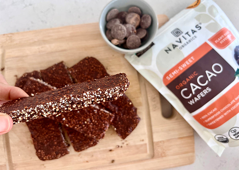 A woman holding a homemade chocolate crisp bar made with Navitas Organics Semi-sweet Cacao Wafers above a cutting board topped with more bars and cacao wafers