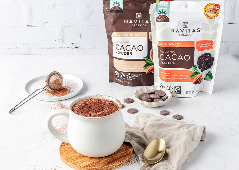 A mug filled with a cacao latte made with Navitas Organics Cacao Powder and Semi-sweet Cacao Wafers in front of bags of the products and a dish filled with cacao wafers
