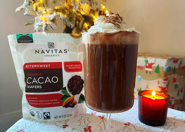 A tall glass filled with a bone broth hot chocolate next to a candle and a bag of Navitas Organics Bittersweet Cacao Wafers, set in a holiday scene