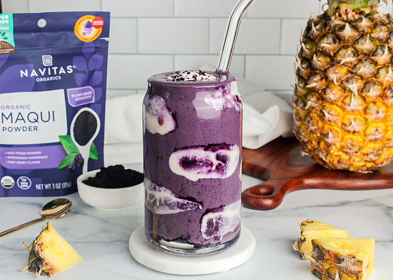 A tall glass filled with a vibrant purple smoothie surrounded by a bag of Navitas Organics Maqui Powder, a fresh pineapple, small pineapple slices and a small dish filled with maqui powder.