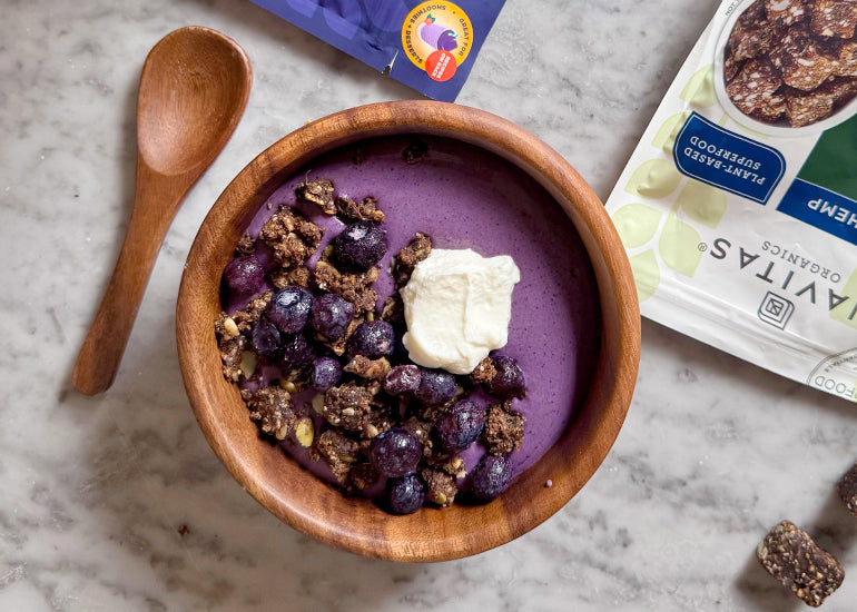 A wooden bowl filled with a maqui berry smoothie topped with fresh blueberries, a dollop of Greek yogurt and crumbled Navitas Organics Blueberry Hemp Power Snacks.