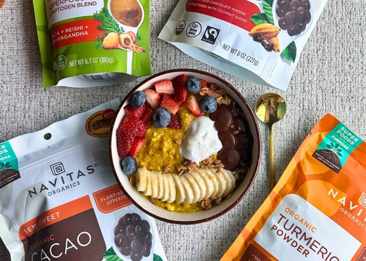 A bowl of oatmeal topped with fresh berries, banana slices, granola and yogurt, surrounded by bags of Navitas Organics Superfood+ Adaptogen Blend, Semi-sweet Cacao Wafers and Turmeric Powder.