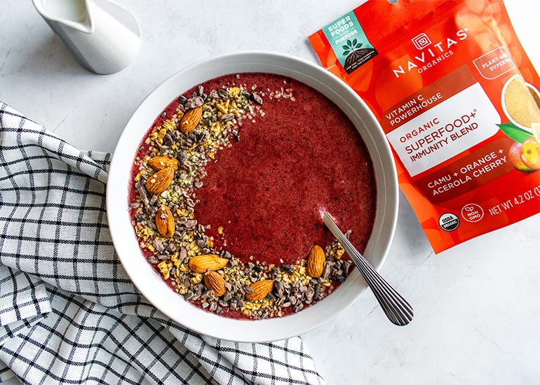 A citrus smoothie bowl topped with flax seeds, chia seeds, hemp seeds and almonds, surrounded by a dish towel, carafe of milk and a bag of Navitas Organics Superfood+ Immunity Blend.