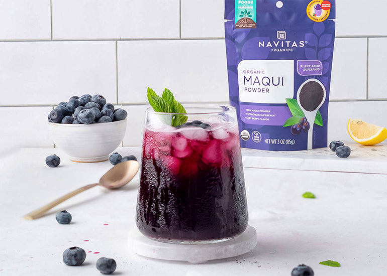 A short glass filled with a purple drink surrounded by fresh blueberries and a package of Navitas Organics Maqui Powder.