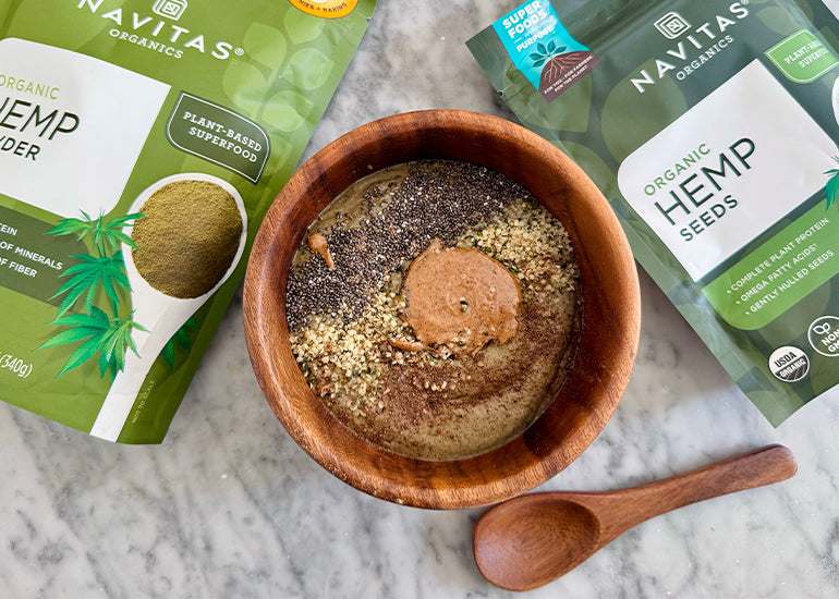 A smoothie bowl in a wooden bowl topped with chia seeds, hemp seeds and nut butter, surrounded by a wooden spoon, a bag of Navitas Organics Hemp Powder and a bag of Navitas Organics Hemp Seeds.