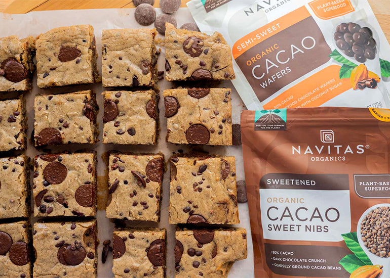 Rows of blondies on parchment paper next to bags of Navitas Organics Semi-sweet Cacao Wafers and Cacao Sweet Nibs.