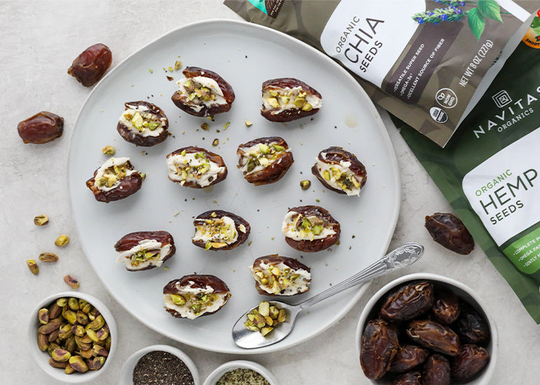 A plate of cream cheese stuffed dates topped with crushed pistachios, hemp seeds and chia seeds, next to bags of Navitas Organics Chia Seeds and Hemp Seeds