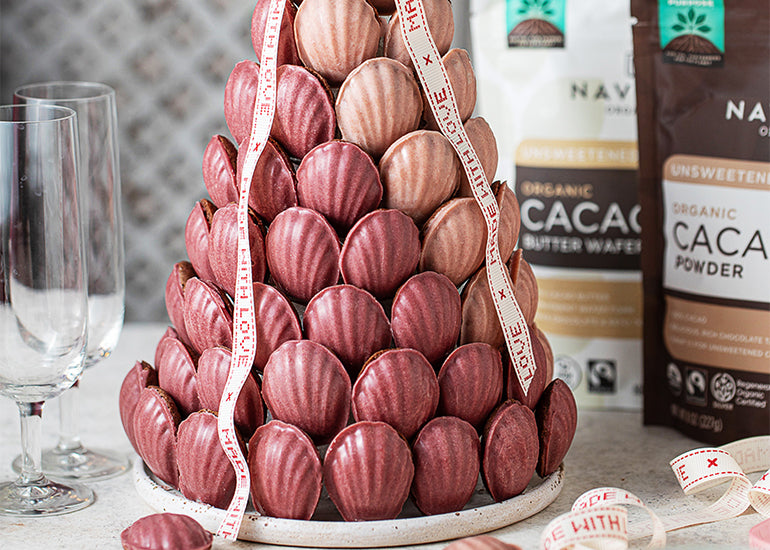 A Valentine's themed madeleine cookie tower beside empty champagne glasses and bags of Navitas Organics Cacao Powder and Cacao Butter Wafers.