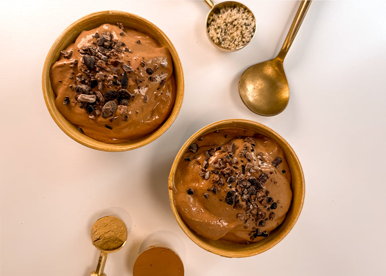 Two bowls of homemade chocolate peanut butter ice cream topped with cacao nibs, surrounded by gold spoons filled with hemp seeds, maca powder and cacao powder.