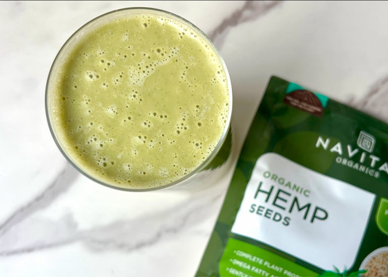 An overhead shot of a creamy green smoothie in a tall glass next to a bag of Navitas Organics Hemp Seeds laying on a marble counter