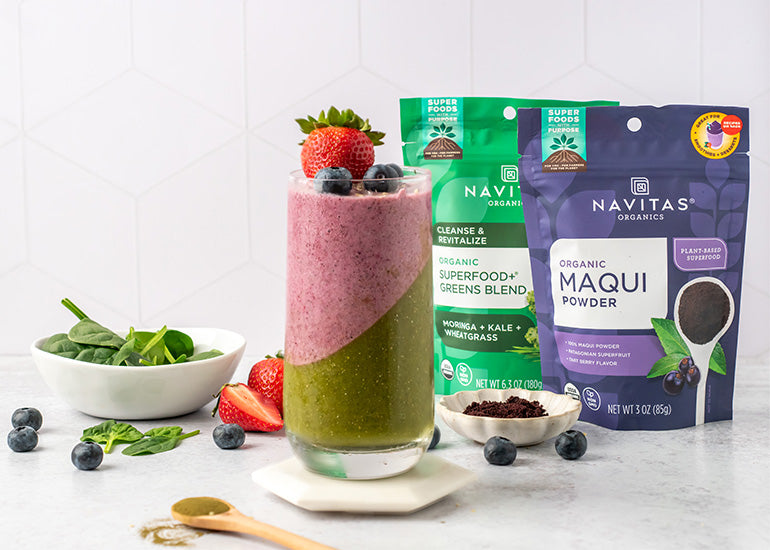 A tall layered greens and berry smoothie topped with a fresh strawberry and blueberries, surrounded by fresh spinach leaves and bags of Navitas Organics Superfood+ Greens Blend and Maqui Powder.