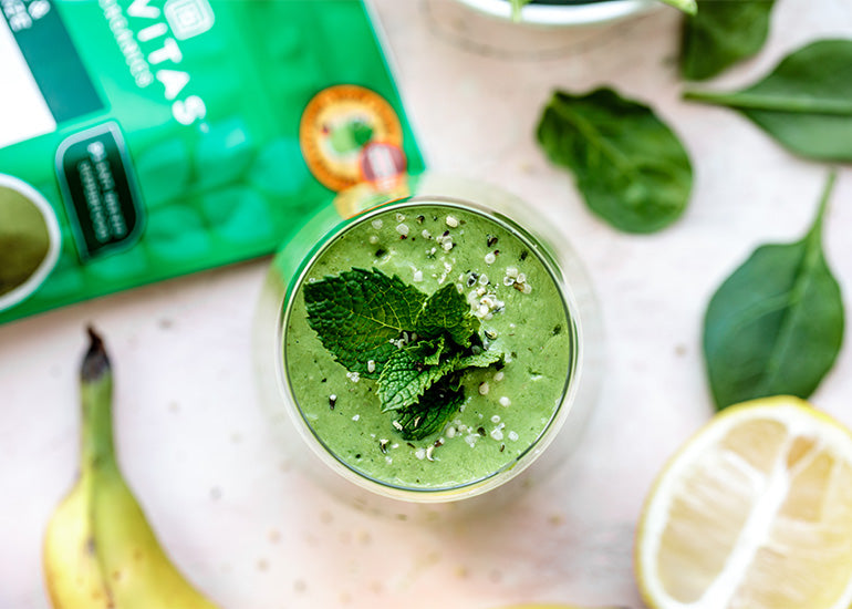 An overhead shot of a green smoothie topped with fresh mint leaves and hemp seeds surrounded by a banana, lemon, spinach leaves and a bag of Navitas Organics Superfood+ Greens Blend.