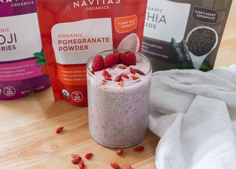 A glass dish filled with a pomegranate chia pudding topped with fresh raspberries and goji berries surrounded by bags of Navitas Organics Goji Berries, Pomegranate Powder and Chia Seeds