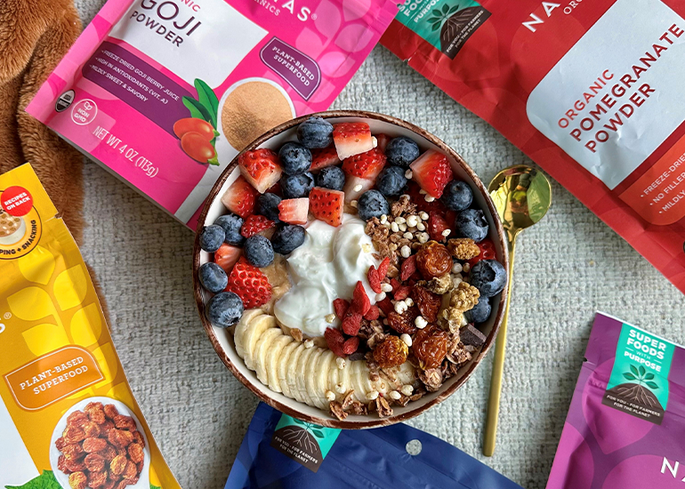 A bowl of oats mixed with Navitas Organics Pomegranate Powder, topped with fresh berries, banana slices, Greek yogurt, and a variety of Navitas Organics superfood berries, surrounded by bags of Navitas Organics superfood products.