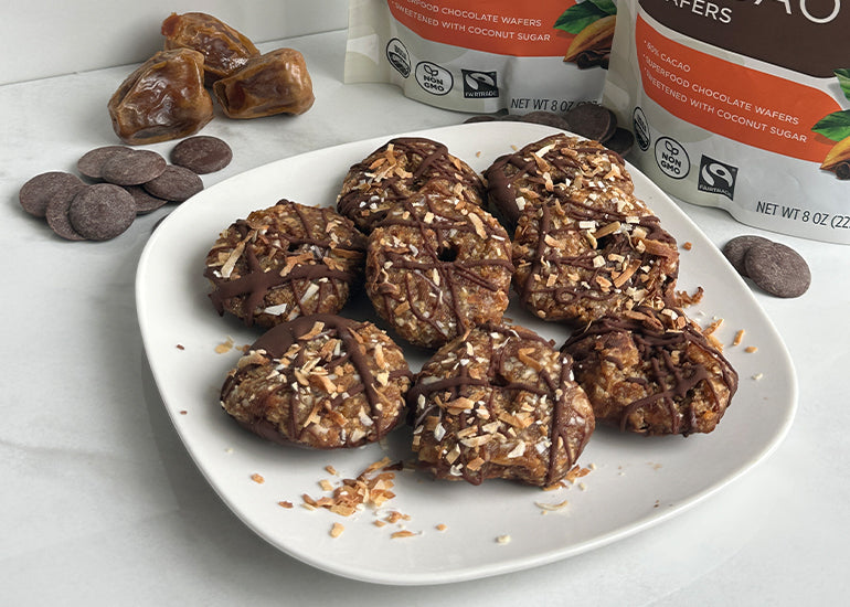 A plate of no-bake chocolate coconut cookies surrounded by piles and bags of Navitas Organics Semi-sweet Cacao Wafers and dates.