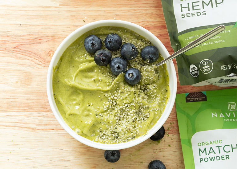 A green smoothie bowl topped with hemp seeds and blueberries on a wooden surface surrounded by scattered blueberries and bags of Navitas Organics Hemp Seeds and Matcha Powder