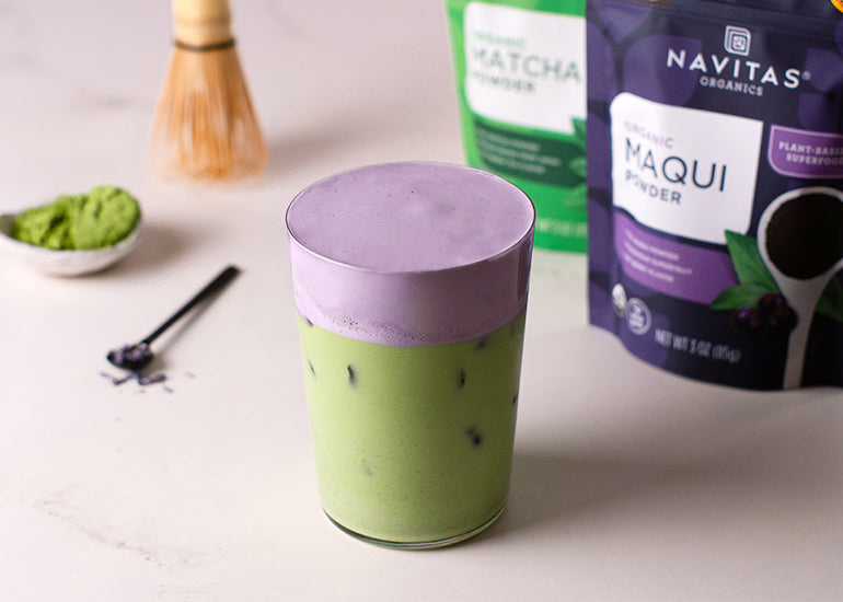 An iced matcha latte in a tall glass topped with purple cold foam cream next to a dish filled with matcha powder, a matcha whisk, and bags of Navitas Organics Matcha Powder and Maqui Powder.