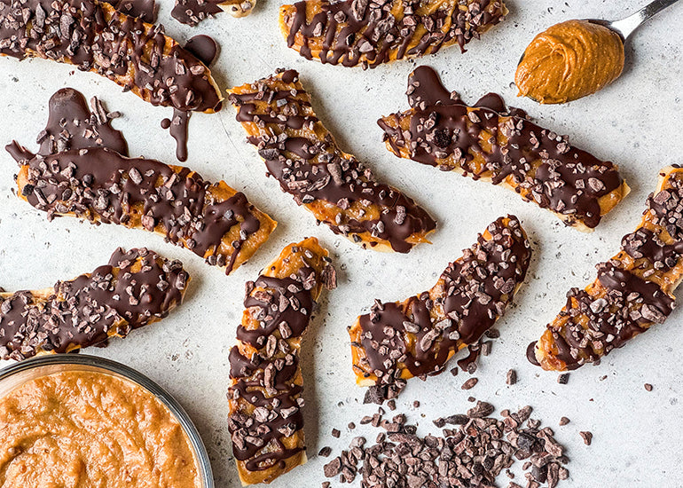 An overhead shot of frozen cacao banana bars layered with peanut butter, a date caramel sauce, and cacao nibs, surrounded by a spoonful and a dish full of peanut butter, along with a small pile of cacao nibs.