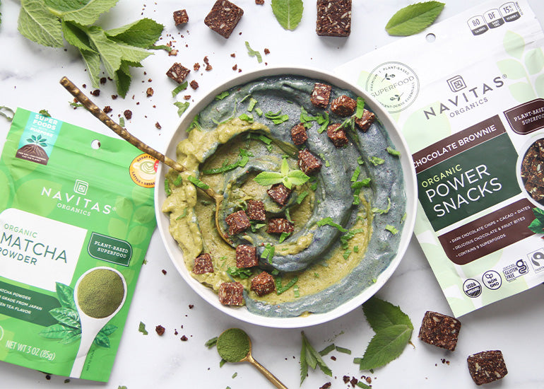 A green and blue swirled smoothie bowl topped with chopped Navitas Organics Chocolate Brownie Power Snacks, surrounded by bags of Navitas Organics Matcha Powder and Chocolate Brownie Power Snacks, as well as mint leaves and scattered Power Snacks.